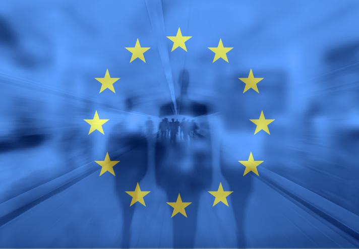 The image shows silhouettes of few persons against a blurred European Union flag, highlighting the circle of twelve gold stars.